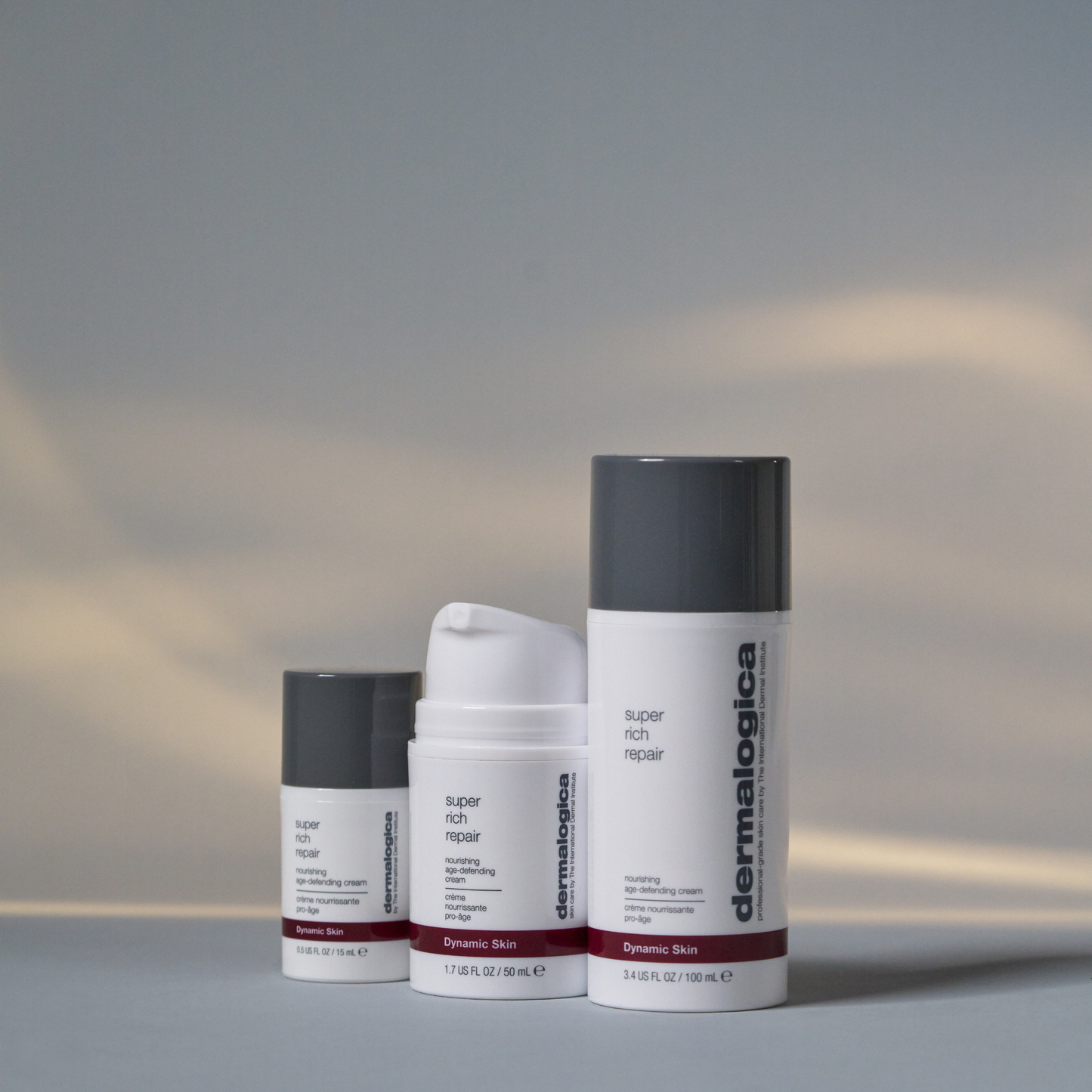 Three Dermalogica super rich repair moisturizing cream bottles in different sizes are displayed side by side on a neutral surface, set against a softly blurred backdrop of light and shadow.