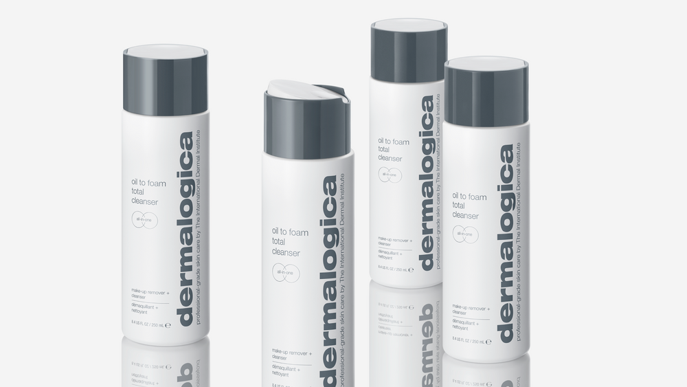 Four bottles of Dermalogica Oil to Foam Total Cleanser are arranged on a white surface, featuring sleek white packaging with gray caps and vertical gray branding text.