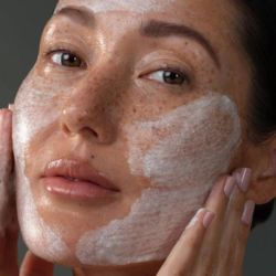 Woman applying a ance mask with a neutral background,  An acne-reducing routine to keep breakouts at bay while caring for the skin underneath.