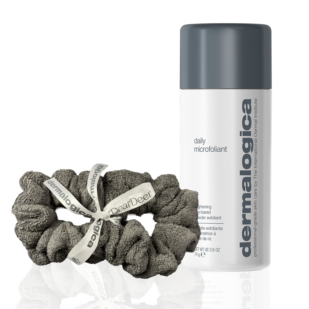 A bottle of Dermalogica daily microfoliant stands upright beside two gray fabric hair scrunchies labeled Dear Deer, all set against a clean white background.