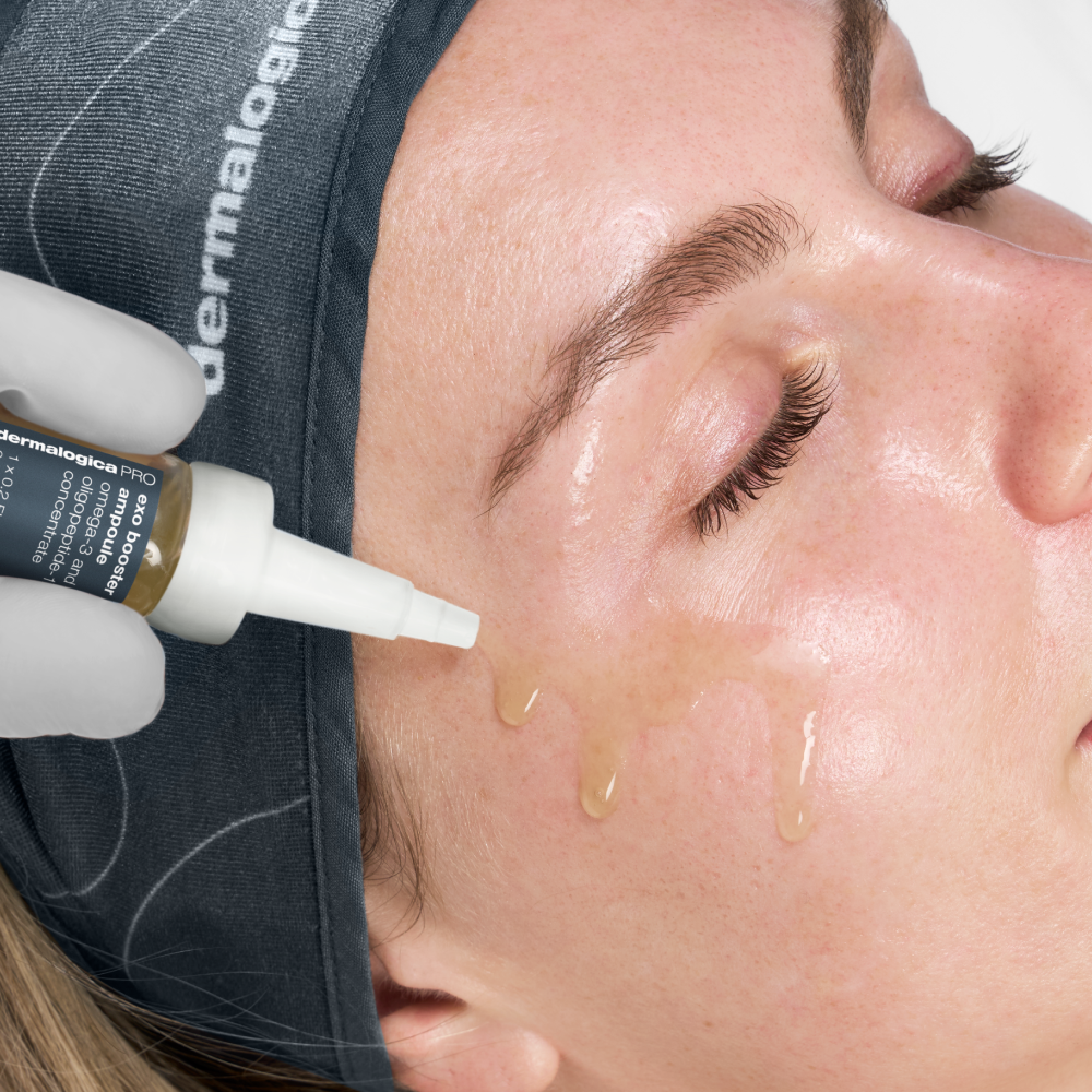 A close-up of a person with closed eyes receiving exo-booster x advanced dermaplaning serum from a bottle held by a gloved hand. They wear a Dermalogica headband, highlighting clean, smoother skin—ideal post-dermaplaning care.