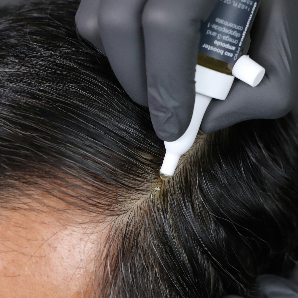 A gloved hand applies Dermalogica PRO exo booster to a client's scalp.