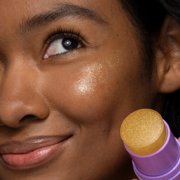 A close-up of a smiling person applying Dermalogica South Africa's golden hour hydrating spf30 stick to their cheekbone, enhancing their glow with a non-comedogenic formula that leaves skin radiant and fresh without clogging pores.