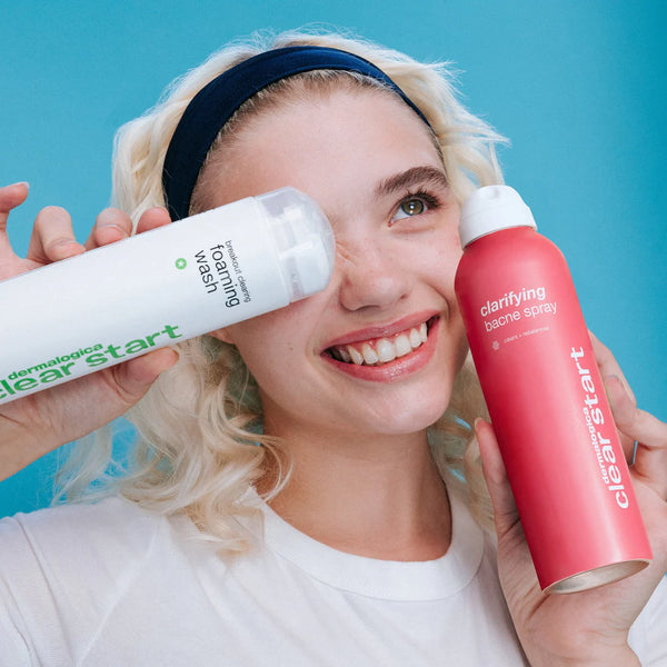 A smiling person with blonde hair holds the Dermalogica South Africa breakout rescue trio—a white foaming wash bottle and a pink bacne spray—against a blue background, perfect for treating body acne.