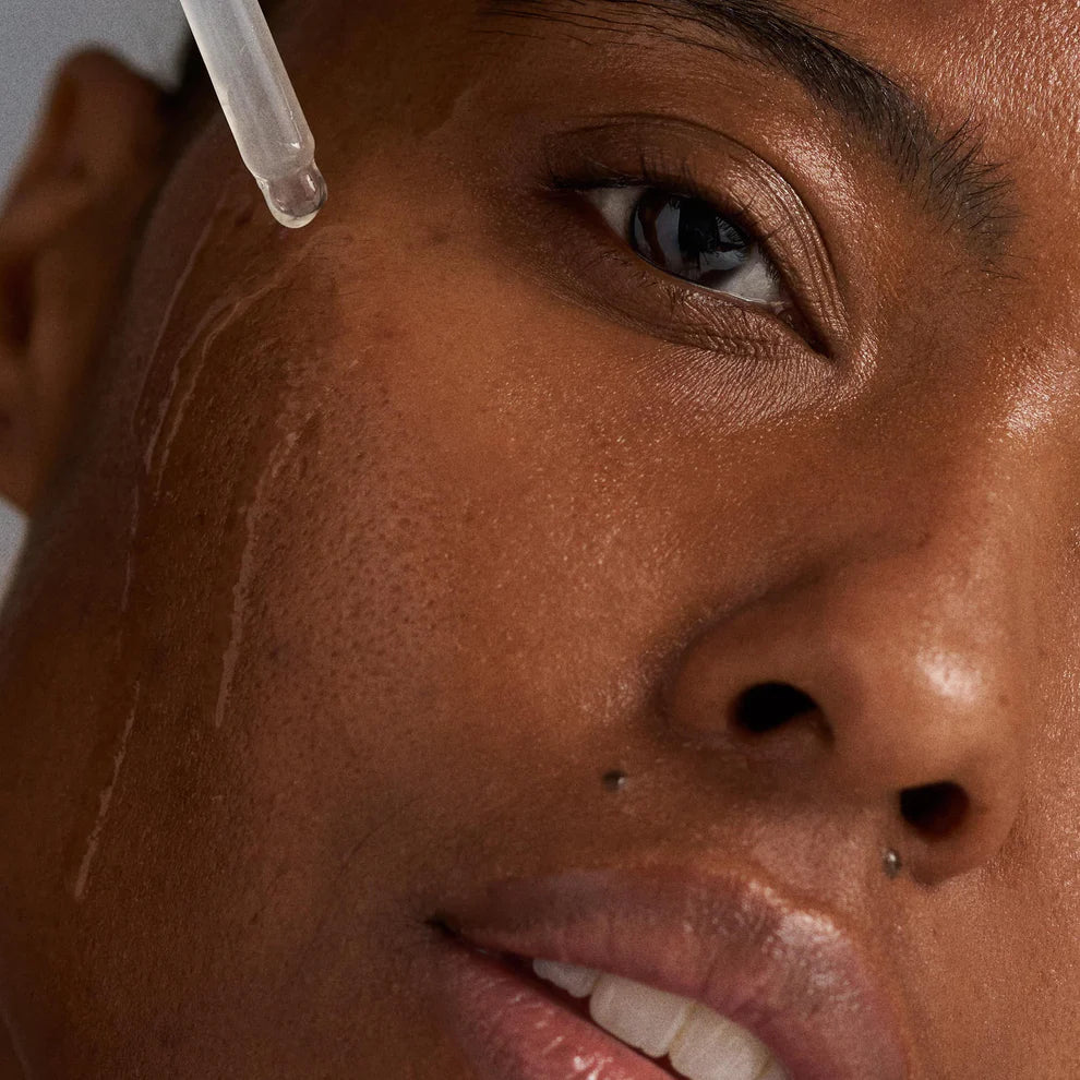 A close-up of a person with smooth brown skin and a nose piercing, applying Dermalogica South Africa's hydration you can feel kit (2 full-size) serum from a dropper to their cheek, highlighting visible pores and deep hydration.