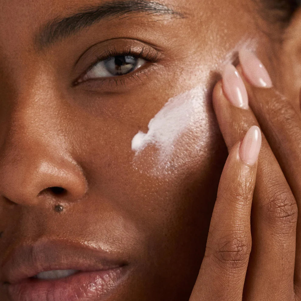 A close-up of a person with medium-dark skin applying Dermalogica South Africa’s get + keep your glow kit (2 full-size) to their cheek using their fingertips; their eye, lips, and neatly manicured nails are visible.