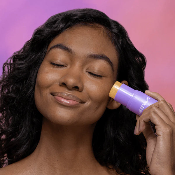 A woman with curly hair smiles with her eyes closed while applying Dermalogica South Africa's golden hour hydrating spf30 stick, a non-comedogenic SPF stick in a purple container, against a pink and purple gradient background.
