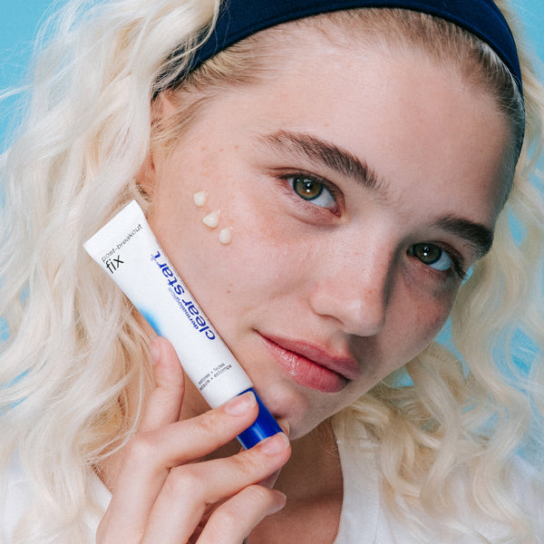 A young person with light skin and blonde hair applies three dots of Dermalogica post-breakout fix to their cheek, holding the tube against a blue background.