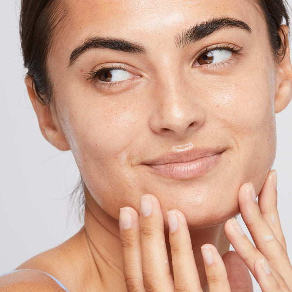 A woman with glowing, smooth skin smiles softly while touching her face, looking content. Thanks to Dermalogica's calm water gel with hyaluronic acid, her makeup-free look shines as it hydrates her dry skin.