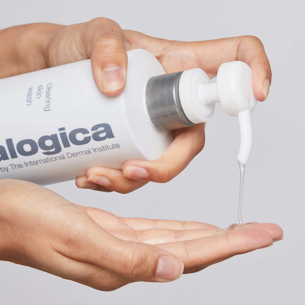 A person dispenses Dermalogica clearing skin wash, a clear gel foaming cleanser, from a white bottle onto their palm against a neutral background.
