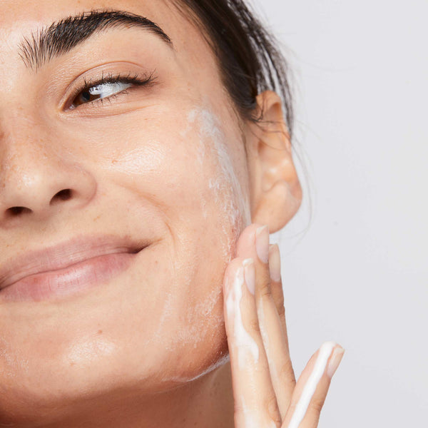 A woman with dewy, healthy skin and dark eyebrows smiles as she applies Dermalogica Clearing Skin Wash foaming cleanser to her cheek with her hand. She has a natural look against a plain light background.