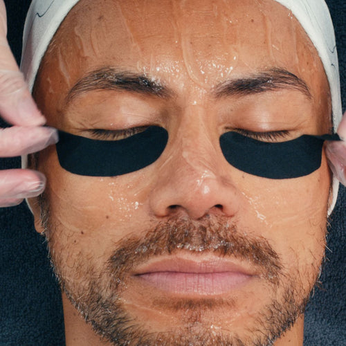 With eyes closed, a person with a towel on their head receives the Dermalogica pro eye flash treatment as gloved hands apply black, botanical-infused under-eye patches over a shiny facial mask.