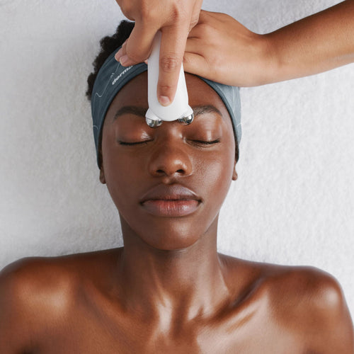 A person with smooth skin and a dark headband relaxes on a white towel as a white facial roller is used on their forehead during the Dermalogica pro bright treatment, addressing pigmentation concerns.