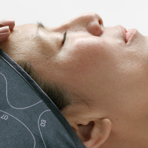 A person relaxes under a gray cloth with white markings, eyes closed, as a hand applies Dermalogica's pro firm treatment to their forehead, delivering a soothing massage that promotes firmer, tighter skin.