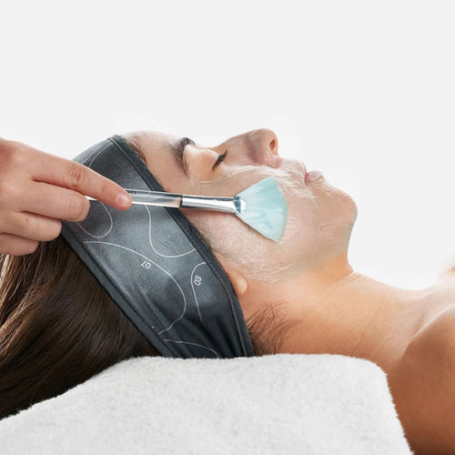 A woman with sensitive skin relaxes on a white towel, wearing a dark headband as Dermalogica's pro calm treatment is gently applied to her face. The clean background highlights this soothing mask that helps strengthen her skin barrier.