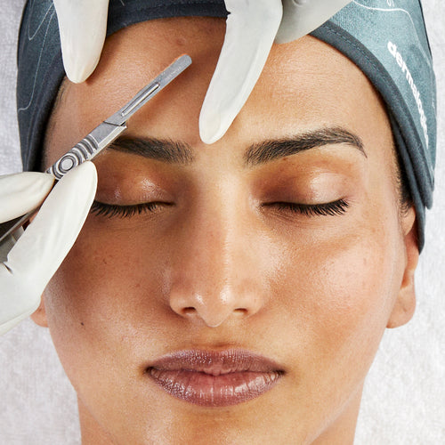 A person with closed eyes receives the Dermalogica pro dermaplaning treatment as a gloved hand uses a surgical blade near their eyebrow to remove dead skin cells. They wear a gray headband and relax on a white surface.