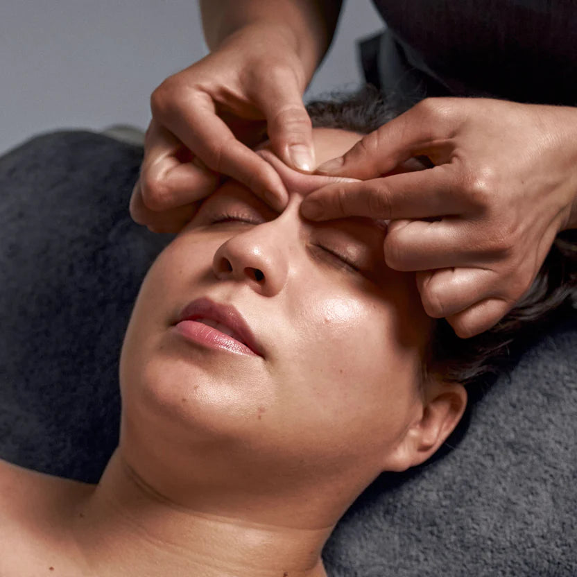 A person relaxes with closed eyes as gentle hands apply the Dermalogica neurosculpt facial treatment, lying on a towel and enjoying the soothing massage for facial sculpting and relaxation.