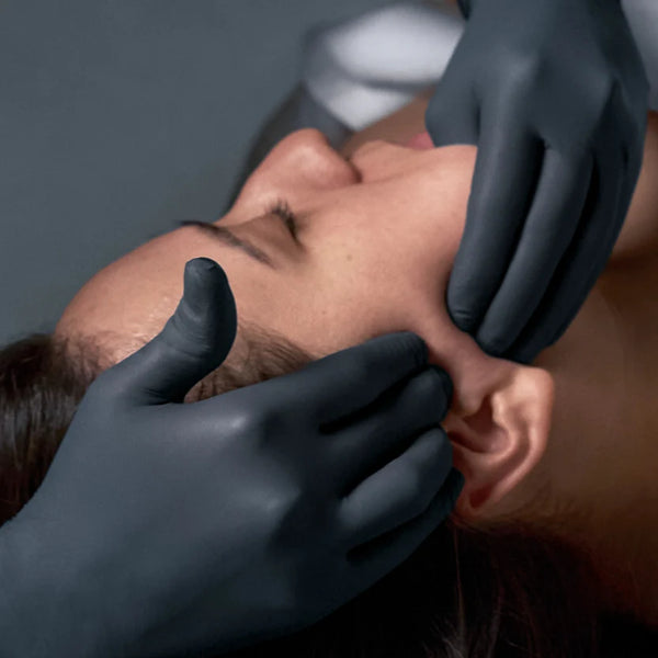 Wearing black gloves, a person gently massages another's jaw and face, demonstrating Dermalogica's neurosculpt massage—an expert facial treatment designed to lift and contour using advanced sculpting techniques.