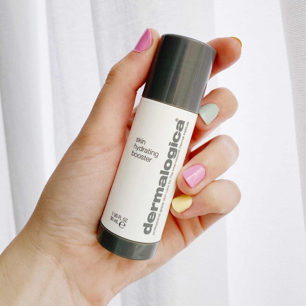 A hand with multicolored pastel nails holds a Dermalogica skin hydrating booster against a light, neutral background for optimal moisture balance.