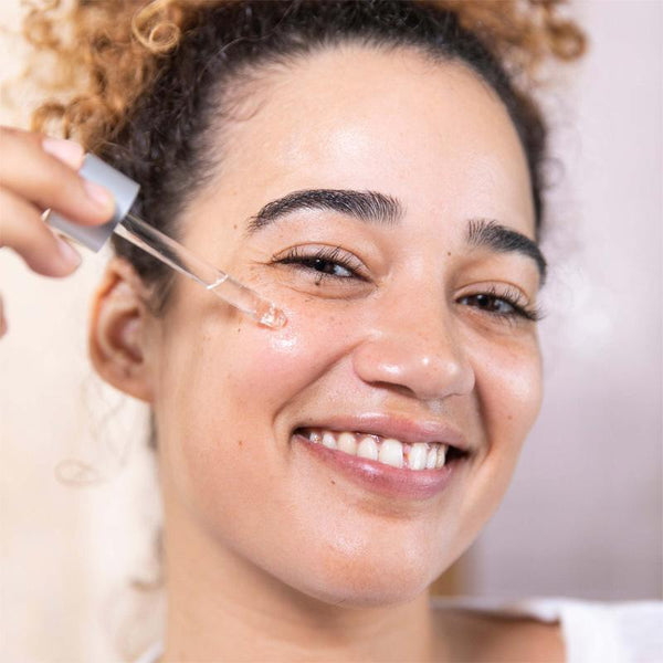 A woman smiles as she applies Dermalogica Age Bright Clearing Serum with a dropper to her cheek, highlighting a salicylic acid-powered skincare routine for clear, glowing skin.