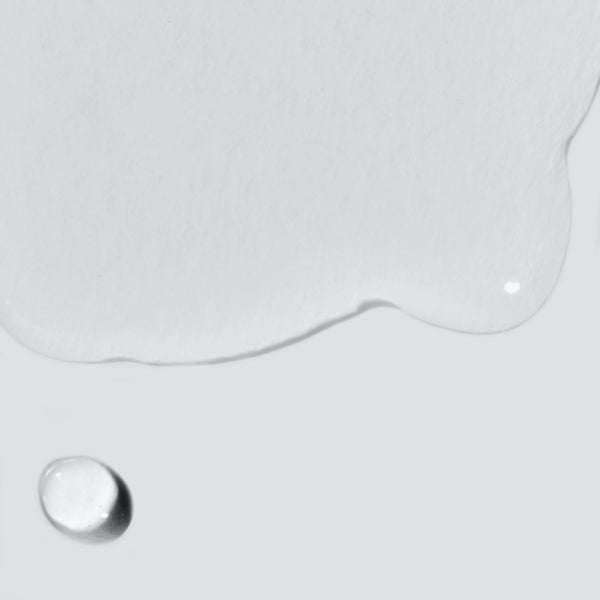 A close-up of Dermalogica Age Bright Clearing Serum with a large irregular salicylic acid puddle and a single droplet on a smooth, light grey surface.