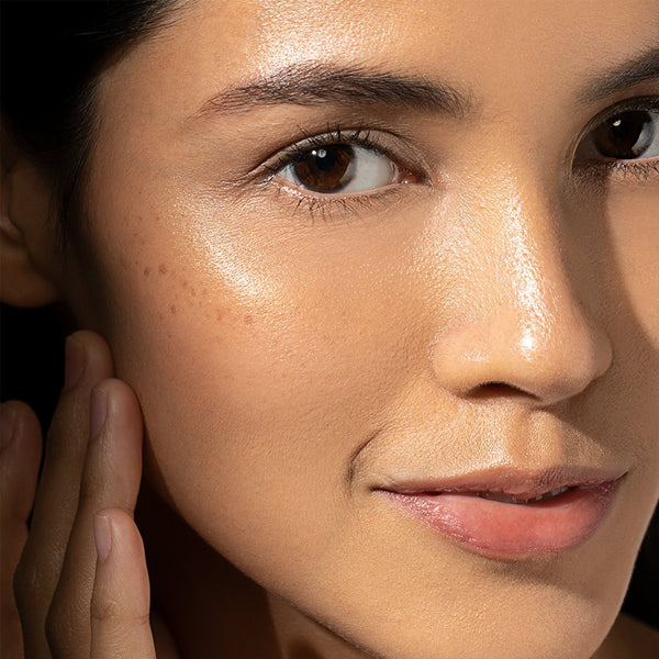 A close-up of a woman with glowing, smooth skin and natural makeup, gently touching her face—her radiant look enhanced by the Dermalogica daily brightness boosters kit.