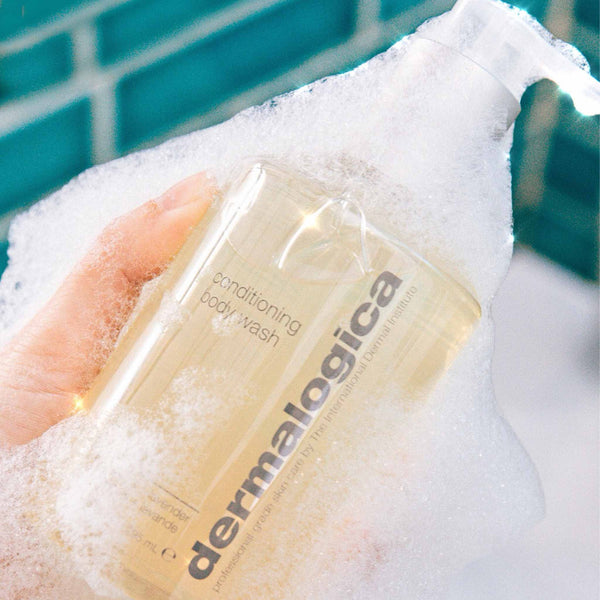 A hand holds a bottle of Dermalogica conditioning body wash, covered in white foam, against a green tiled wall—highlighting its skin-nourishing essential oils blend.