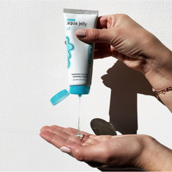 A person squeezes clear gel from a white tube labeled “aqua jelly” with a blue cap onto their palm, against a white background.
